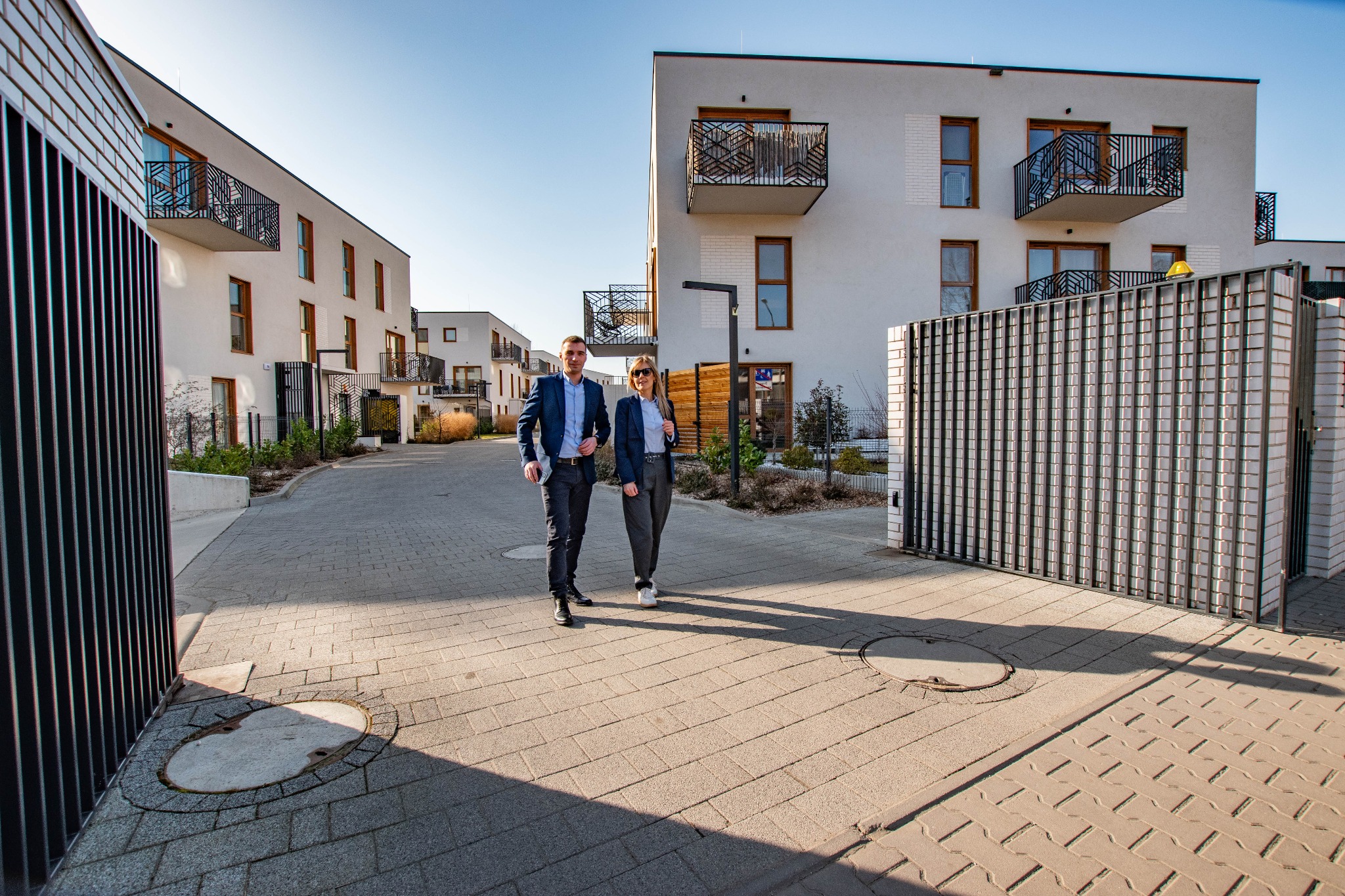 Dwoje agentów nieruchomości w garniturach idzie chodnikiem na tle nowoczesnych budynków mieszkalnych z balkonami i metalowymi ogrodzeniami.