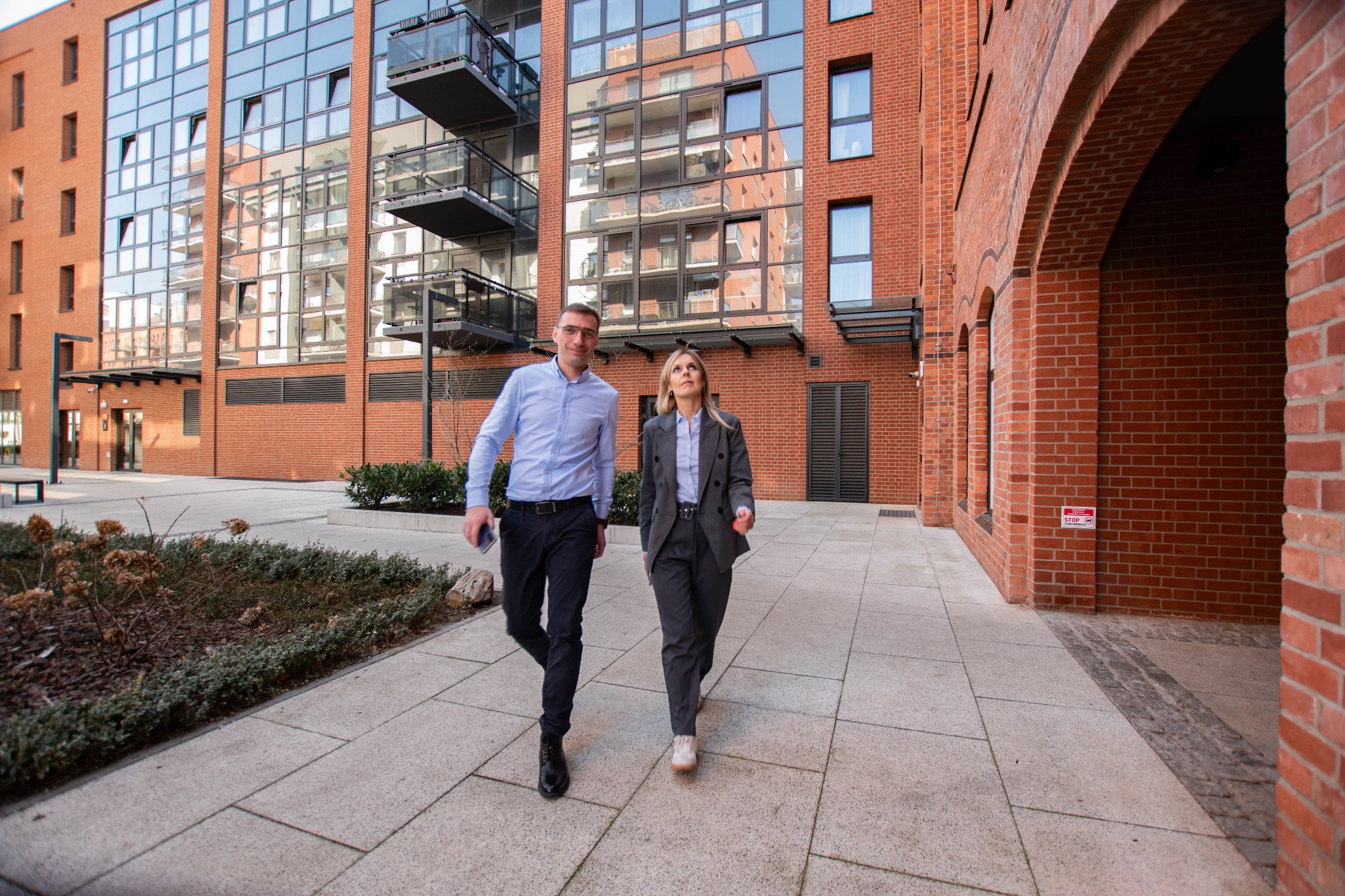 Dwie osoby w strojach biznesowych idą chodnikiem przed nowoczesnym budynkiem z cegły i szkła.
