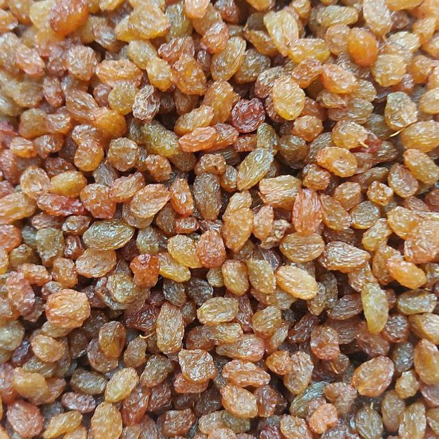 Gęsto ułożone, brązowo-złote rodzynki, niektóre delikatnie błyszczące, wypełniające całą klatkę, o nierównej fakturze i kształtach, z widocznymi drobnymi zmarszczkami na powierzchni.