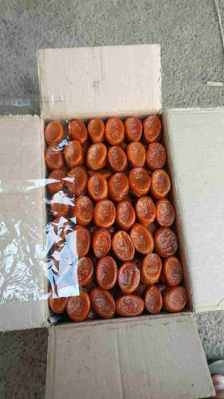 Kartonowe pudełko wypełnione suszonymi morelami, częściowo przykrytymi folią, widoczne na szarym tle.