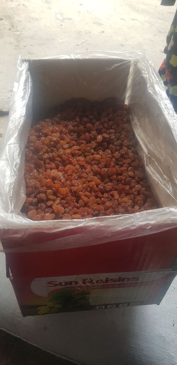 Kartonowe pudełko wypełnione brązowymi rodzynkami, zabezpieczone przezroczystą folią. Widoczny fragment opakowania z napisem 'Sun Raisins'.