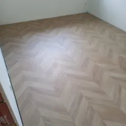 Jasne panele ułożone we wzór jodełki w pustym pokoju, widoczne niedokończone wykończenia przy ścianie.