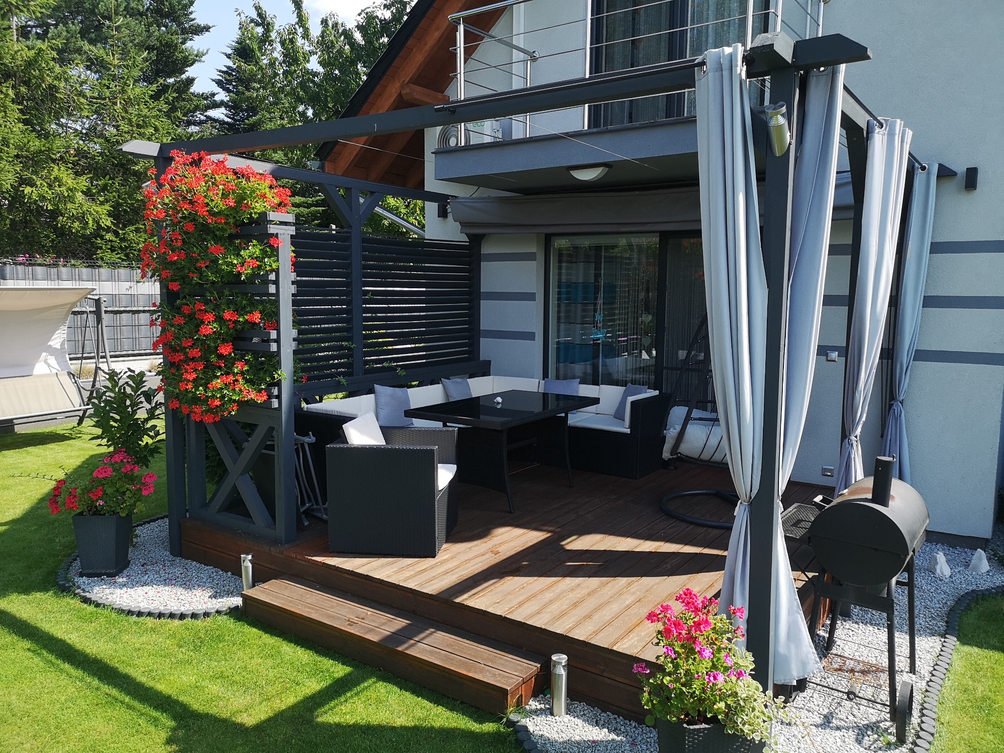 Zadaszony taras z drewnianą podłogą, czarnymi meblami ogrodowymi i pergolą z zasłonami, ozdobiony kwitnącymi pelargoniami w donicach i pnących się po konstrukcji, grill na kółkach z boku.