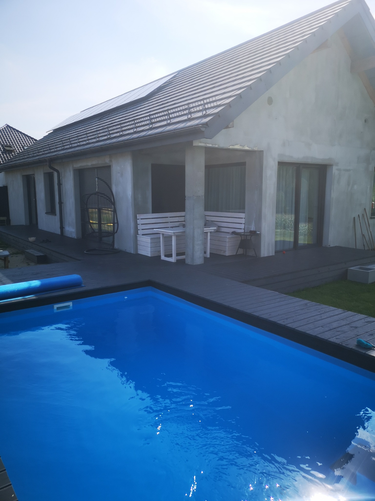 Nowoczesny dom z basenem i ciemnoszarym tarasem kompozytowym, widok z góry na błękitną wodę i fragment budynku z białymi meblami ogrodowymi.