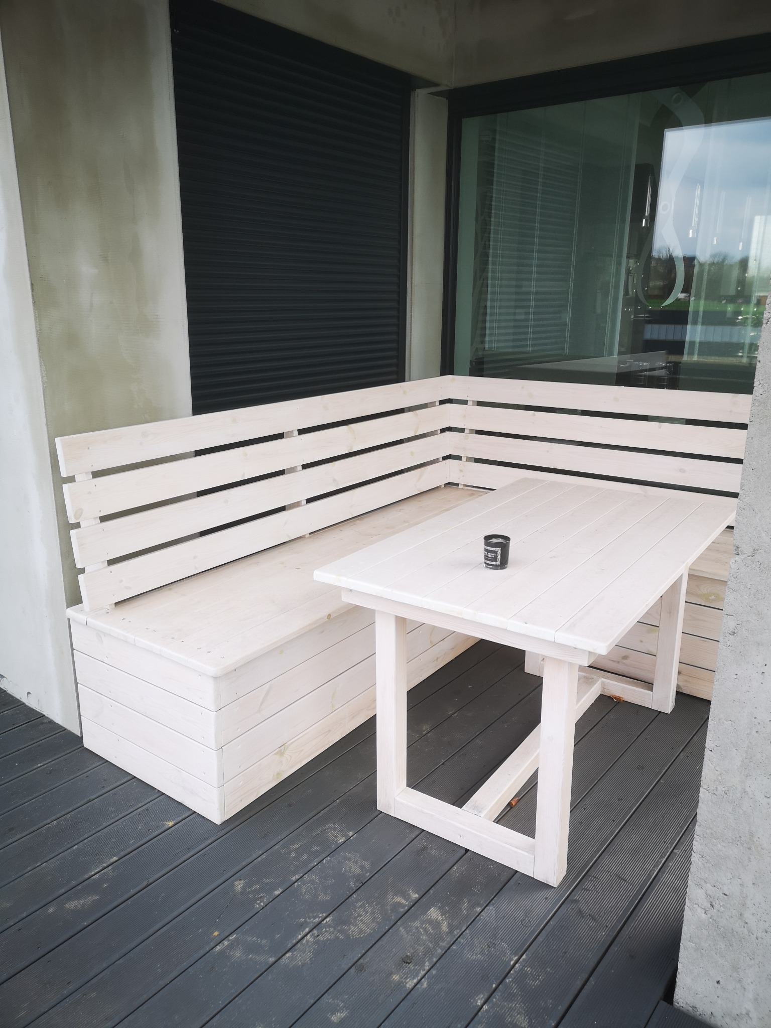 Biała, drewniana ławka w kształcie litery L z oparciem z listew oraz stół z drewna pomalowanego na biało, ustawione na ciemnoszarym tarasie z desek.