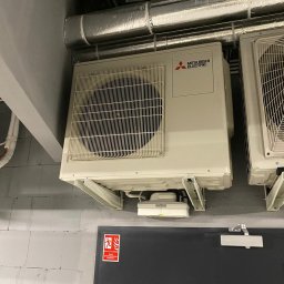 Top Klima - Zamontowane jednostki zewnętrzne klimatyzacji Mitsubishi Electric na szarej ścianie z bloków, widoczne rury wentylacyjne pod sufitem i czarne drzwi z oznaczeniem wyjścia ewakuacyjnego.