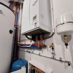 Top Klima - Kotłownia z piecem gazowym Ecodan, zasobnikiem ciepłej wody i rozdzielaczem ogrzewania podłogowego, widoczne rury miedziane, plastikowe i zawory, na ścianie biała farba.