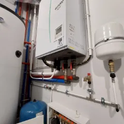 Kotłownia z piecem gazowym Ecodan, zasobnikiem ciepłej wody i rozdzielaczem ogrzewania podłogowego, widoczne rury miedziane, plastikowe i zawory, na ścianie biała farba.