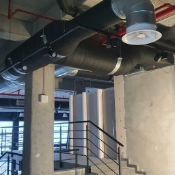 Top Klima - Surowy betonowy strop z widocznymi instalacjami wentylacyjnymi i klimatyzacyjnymi w budynku komercyjnym, schody w tle.