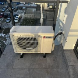 Top Klima - Zewnętrzna jednostka klimatyzacji Mitsubishi Electric na balkonie z widokiem na parking i budynki w tle.