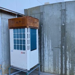 Top Klima - Nowa jednostka zewnętrzna klimatyzacji Mitsubishi Electric, zabezpieczona kartonem na górze, zamontowana na metalowej ramie, stojąca przy betonowej ścianie budynku.