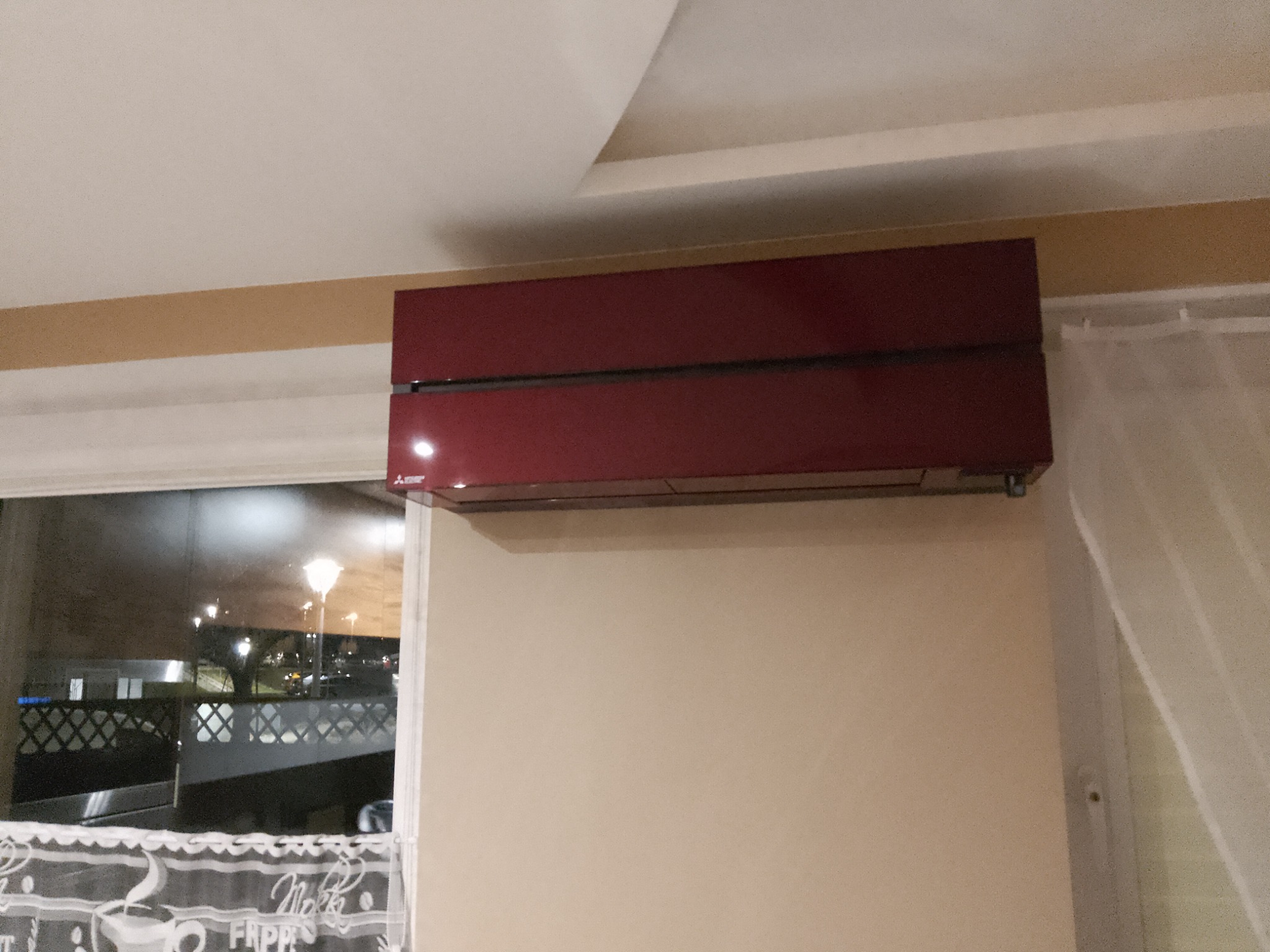 Czerwony klimatyzator ścienny Mitsubishi Electric zamontowany w nowoczesnym wnętrzu, widok z okna na oświetlony parking.