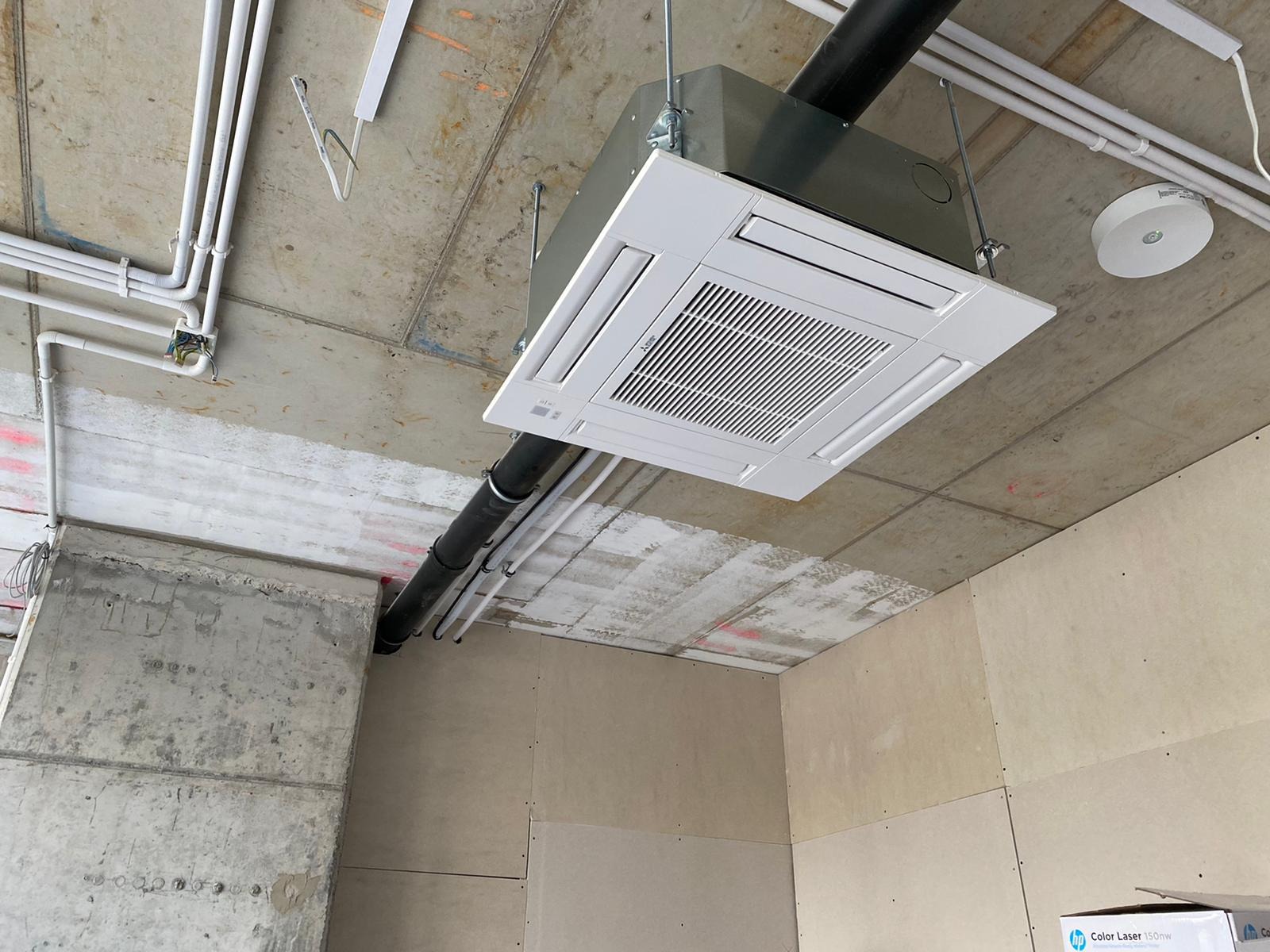 Montaż sufitowej kasety klimatyzacyjnej w surowym wnętrzu, widoczne rury instalacyjne i nieotynkowane ściany.