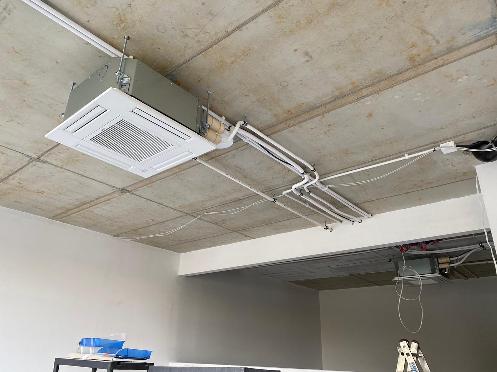 Kasetonowa klimatyzacja sufitowa w surowym wnętrzu, widoczne rury instalacyjne i betonowy strop, drabina w tle.