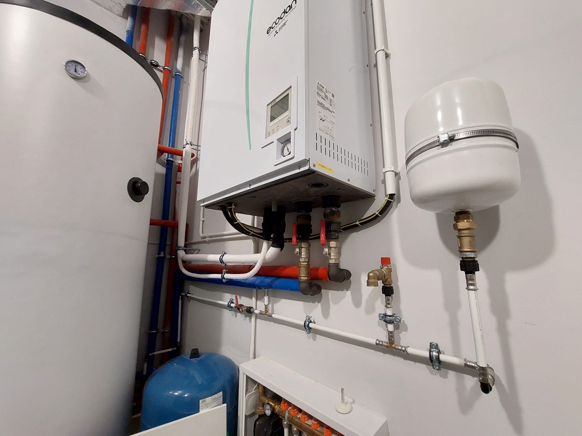 Kotłownia z piecem gazowym Ecodan, zasobnikiem ciepłej wody i rozdzielaczem ogrzewania podłogowego, widoczne rury miedziane, plastikowe i zawory, na ścianie biała farba.