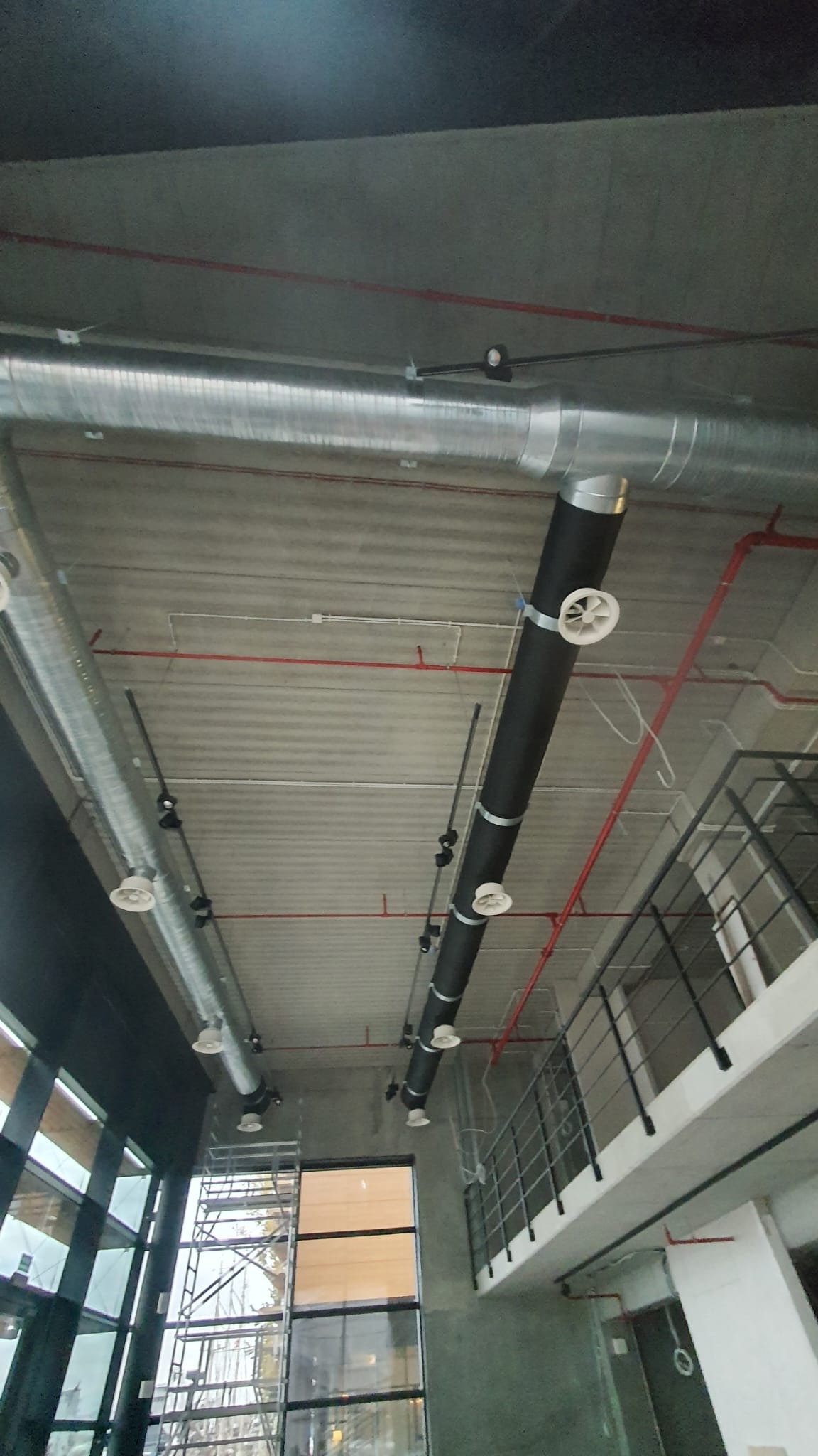 Wnętrze budynku komercyjnego w trakcie instalacji systemu wentylacji: widoczne srebrne i czarne rury wentylacyjne pod sufitem, z białymi anemostatami, na tle surowego betonu i okien z rusztowaniem.
