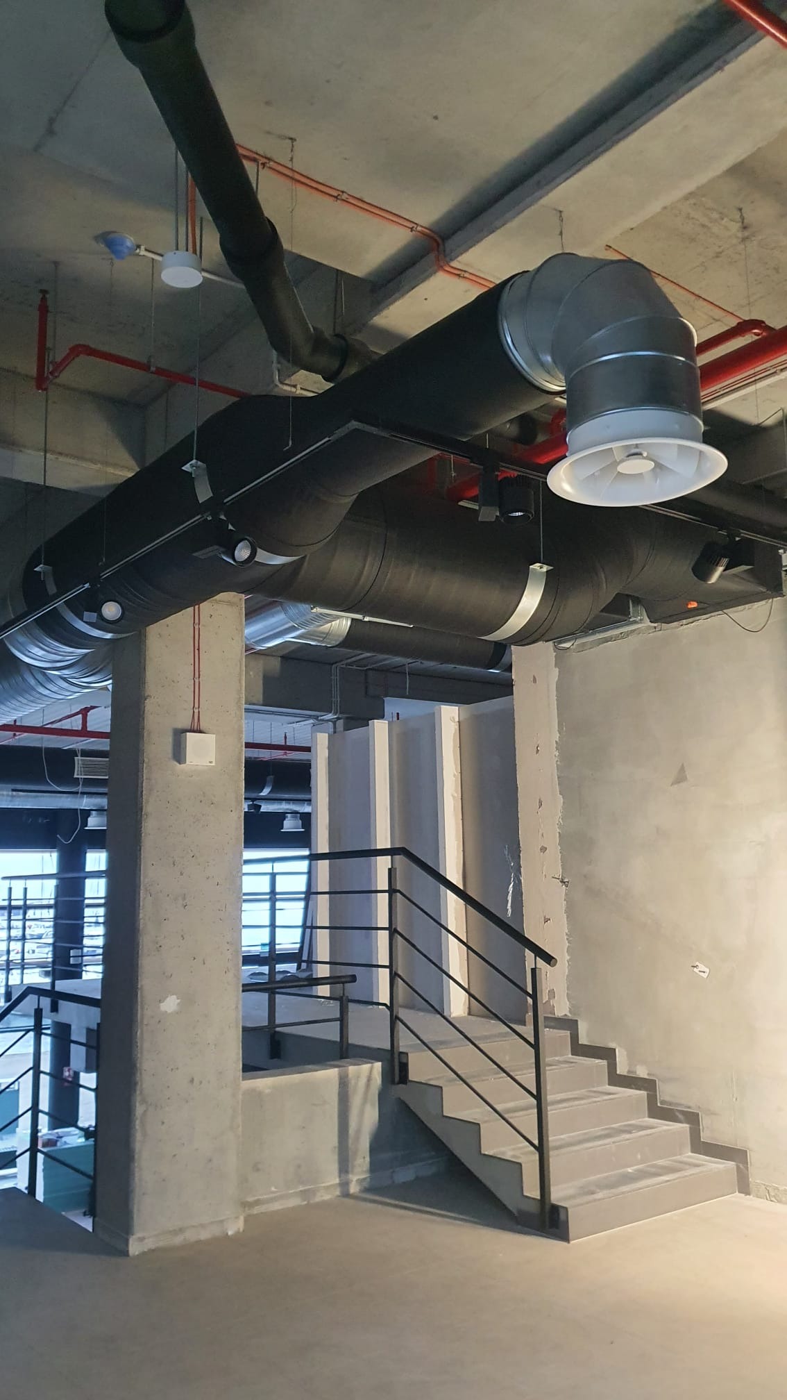 Surowy betonowy strop z widocznymi instalacjami wentylacyjnymi i klimatyzacyjnymi w budynku komercyjnym, schody w tle.