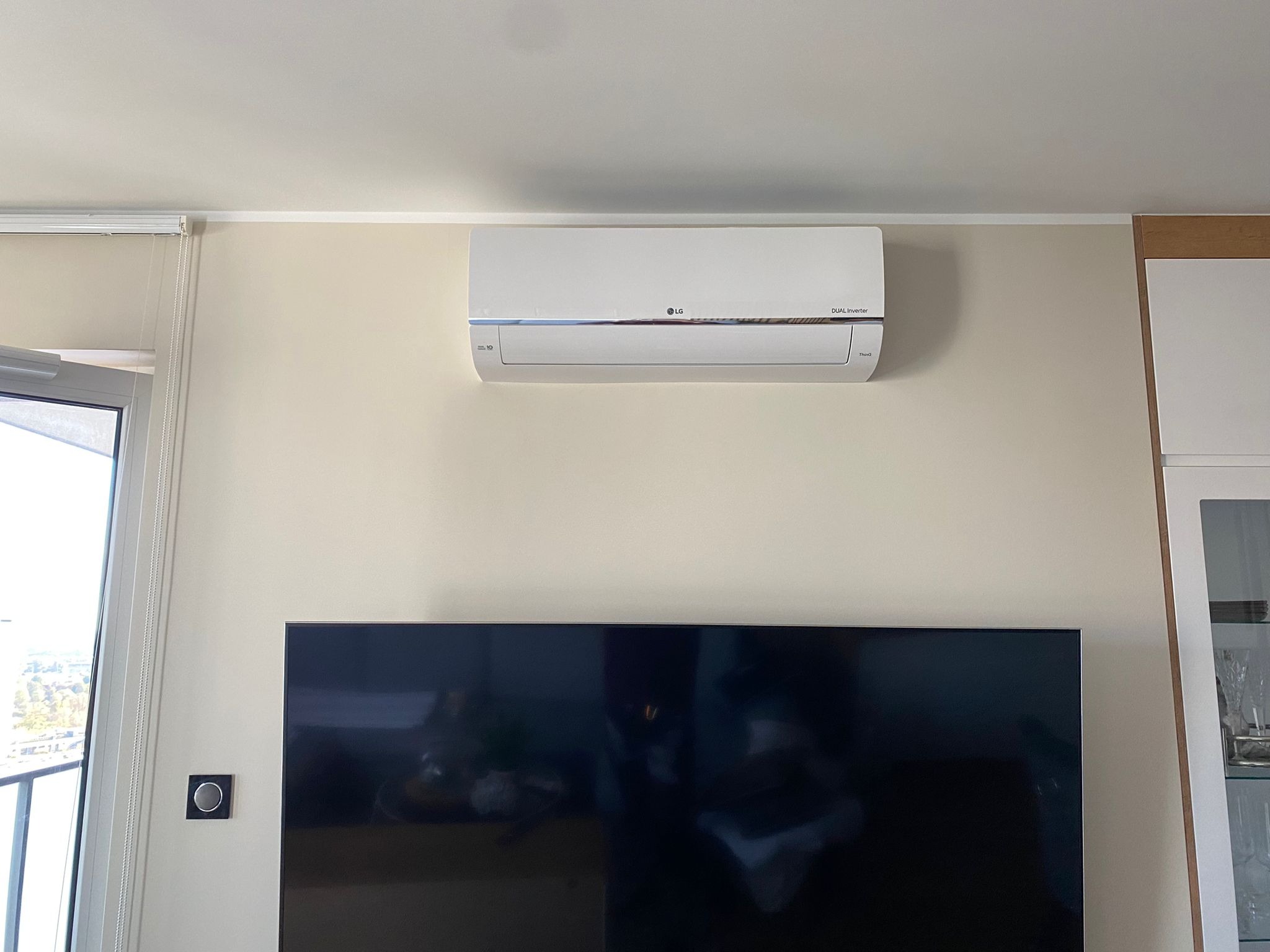 Biały klimatyzator LG Dual Inverter zamontowany na ścianie nad telewizorem w nowoczesnym salonie.