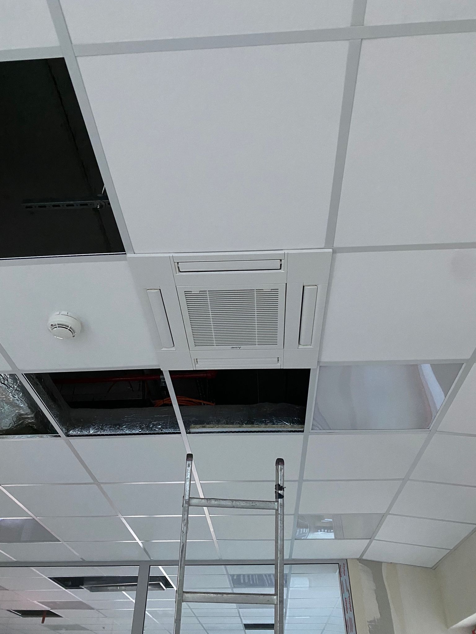 Montaż kasetonowej jednostki klimatyzacji w suficie podwieszanym, widoczna drabina i otwory w suficie z instalacjami.