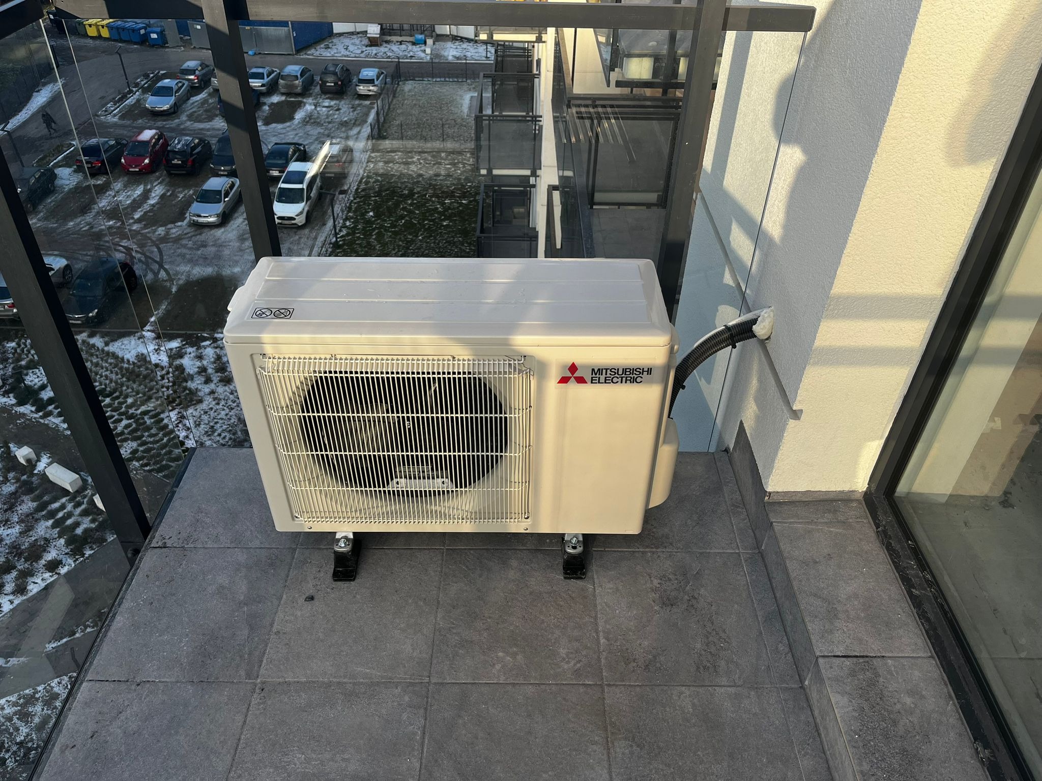 Zewnętrzna jednostka klimatyzacji Mitsubishi Electric na balkonie z widokiem na parking i budynki w tle.