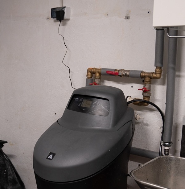 Szary zmiękczacz wody podłączony do instalacji hydraulicznej z zaworami kulowymi i rurami izolowanymi termicznie, widoczne zasilanie elektryczne z gniazdka.