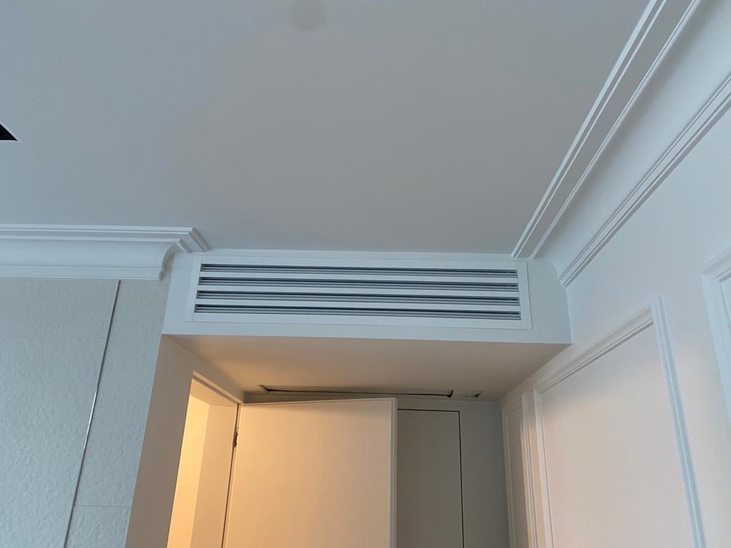 Biała kratka wentylacyjna z poziomymi szczelinami, wkomponowana w sufit z dekoracyjną sztukaterią, widok z dołu na tle otwartych białych drzwi.