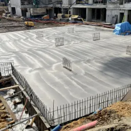 Świeżo wylana, gładka posadzka jastrychowa z wystającymi prętami zbrojeniowymi na placu budowy, widoczne budynki w stanie surowym i sprzęt budowlany w tle.