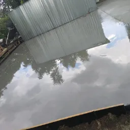 Świeżo wylana, mokra posadzka z widocznym odbiciem nieba i budynku gospodarczego w tle, z użyciem drewnianego szalunku.