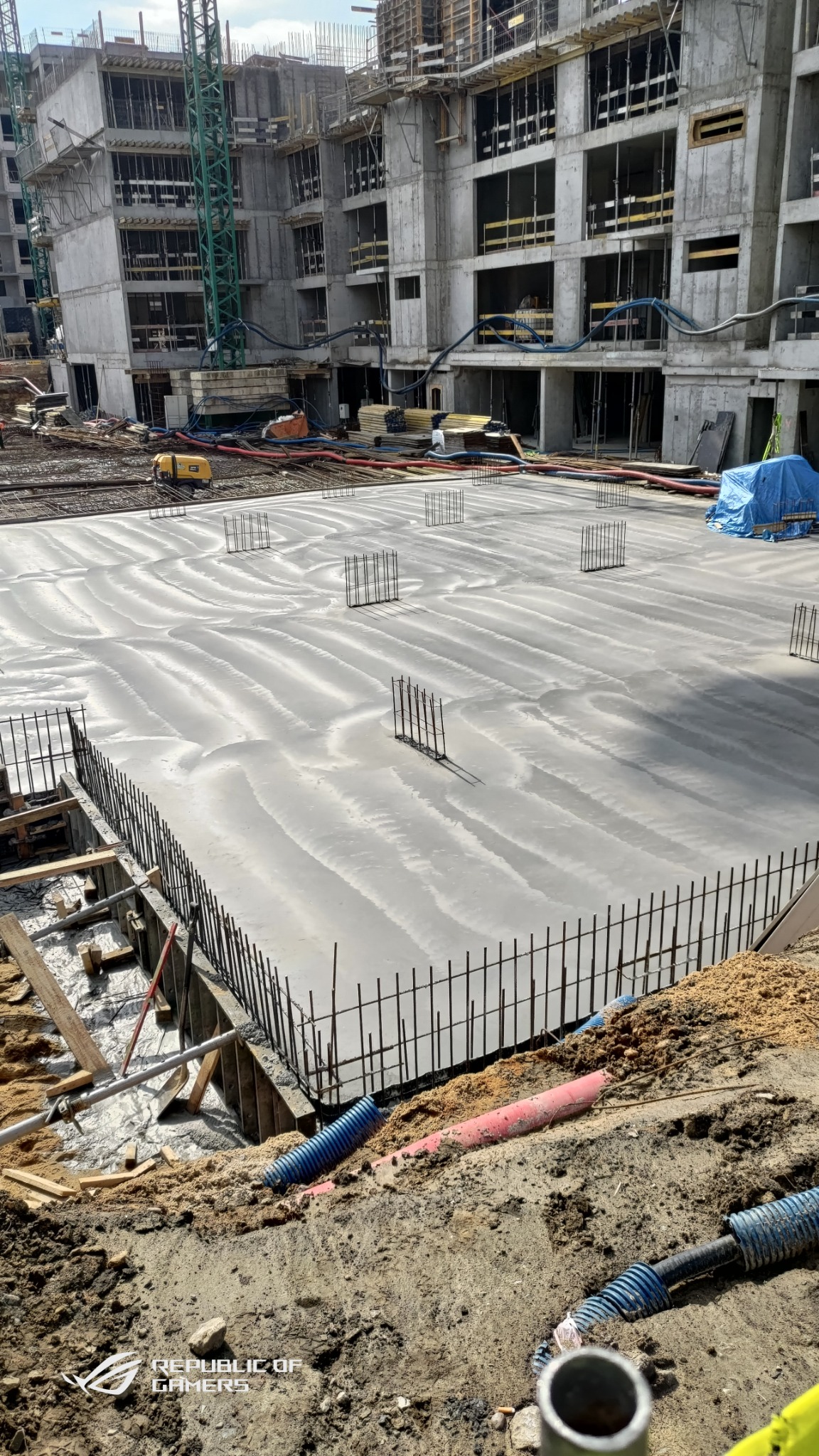 Świeżo wylana, gładka posadzka jastrychowa z wystającymi prętami zbrojeniowymi na placu budowy, widoczne budynki w stanie surowym i sprzęt budowlany w tle.
