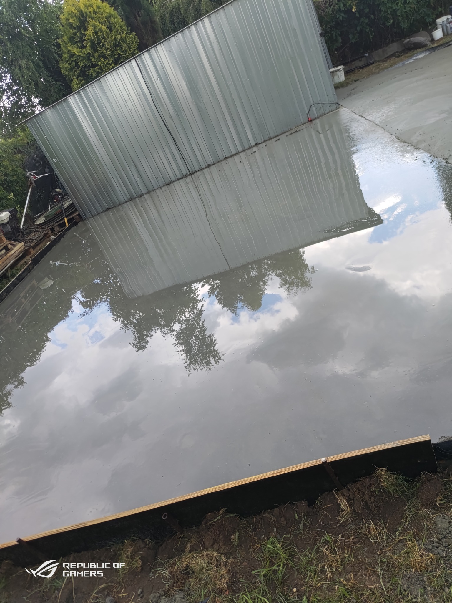 Świeżo wylana, mokra posadzka z widocznym odbiciem nieba i budynku gospodarczego w tle, z użyciem drewnianego szalunku.