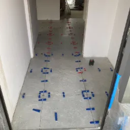 Układanie szarych płytek podłogowych z użyciem poziomicy laserowej i niebieskich oraz czerwonych krzyżyków dystansowych w nowym budynku.