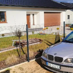 Jasny, parterowy dom z brązową bramą garażową i ciemnym dachem, widziany zza metalowego ogrodzenia i zaparkowanego samochodu BMW, z widocznym cieniem osoby robiącej zdjęcie.