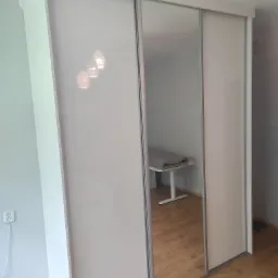 Biała szafa przesuwna z lustrem, odbijająca fragment pokoju z biurkiem i złożonym kocem.