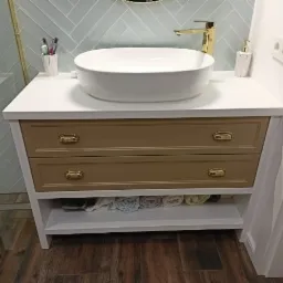 Łazienka z umywalką nablatową na szafce z dwoma szufladami w kolorze taupe i otwartą półką na ręczniki, złota armatura, tło z płytek ułożonych w jodełkę.