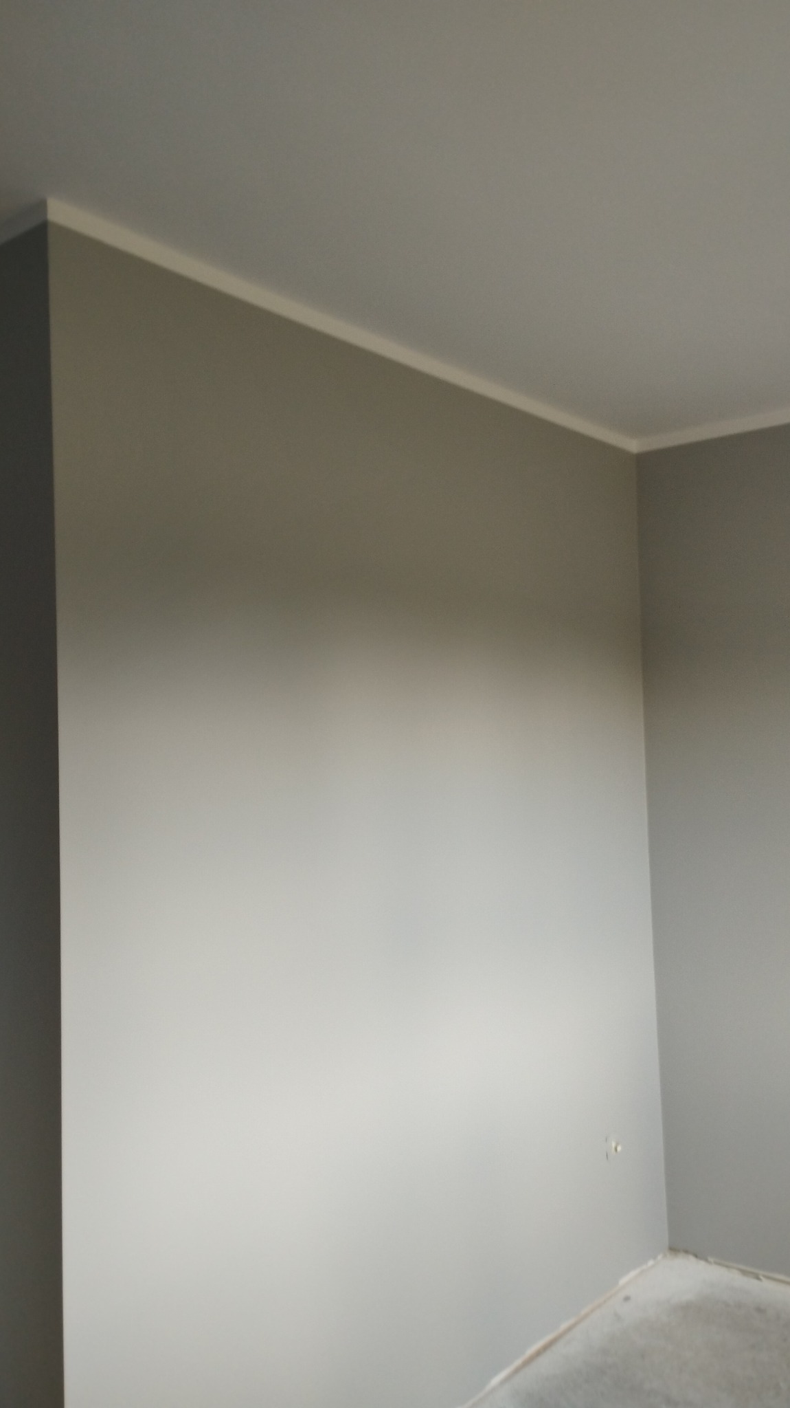 Szara ściana po malowaniu z białą listwą przysufitową, widoczne ślady po taśmie malarskiej na podłodze w rogu pokoju.
