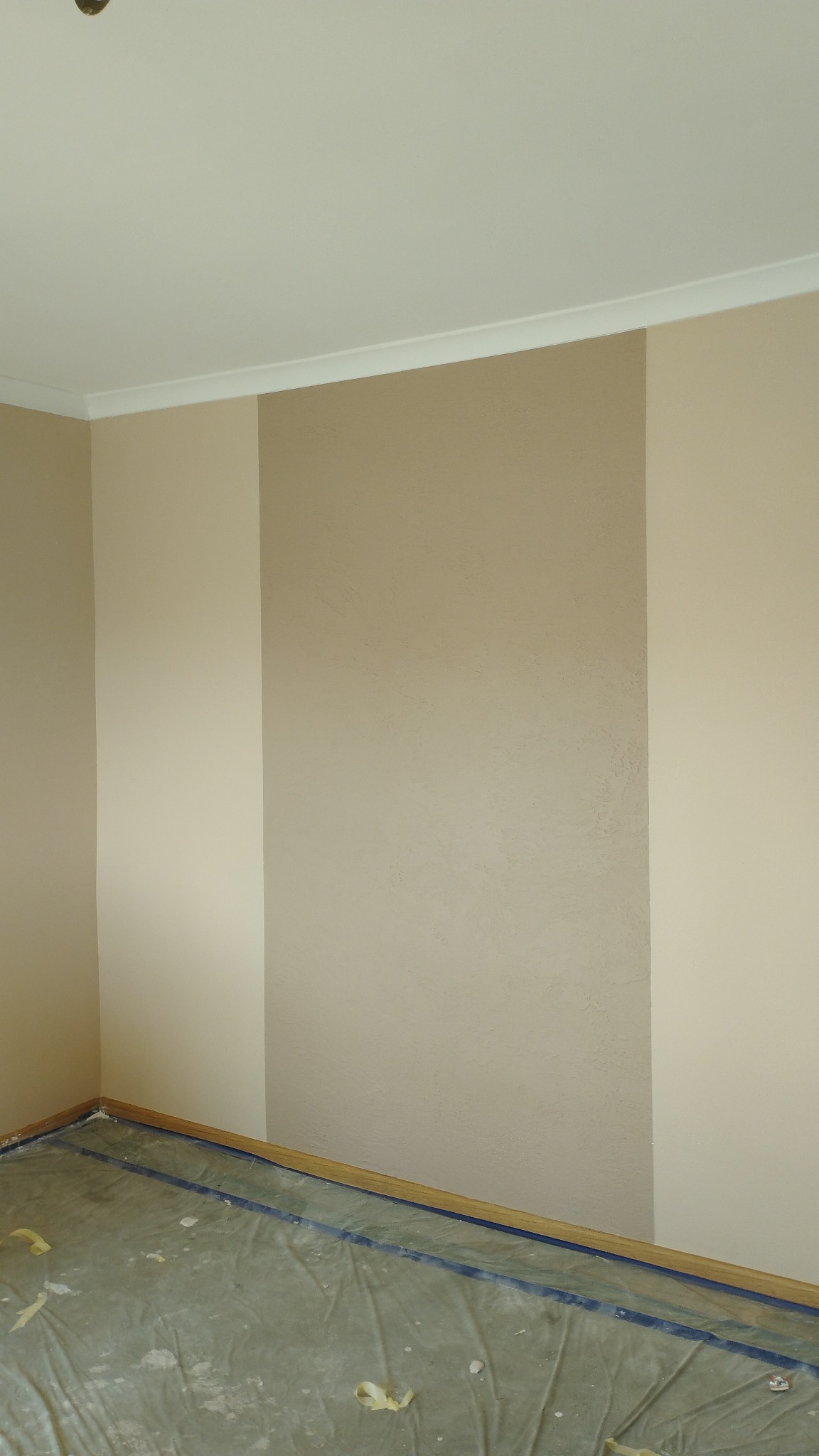 Pomalowane ściany w odcieniach beżu z pionowym pasem o innej fakturze, podłoga zabezpieczona folią malarską.