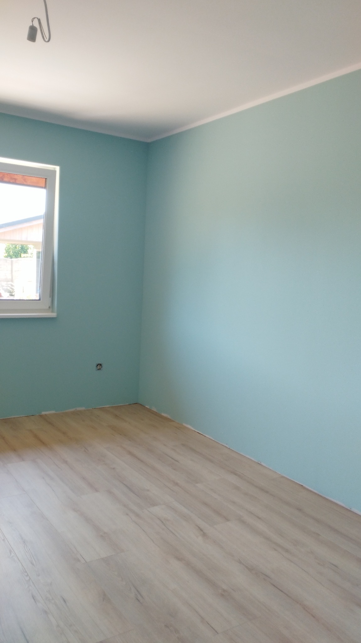 Pomalowane na turkusowo wnętrze pokoju z jasną podłogą laminowaną i białym oknem z widokiem na zieleń na zewnątrz.