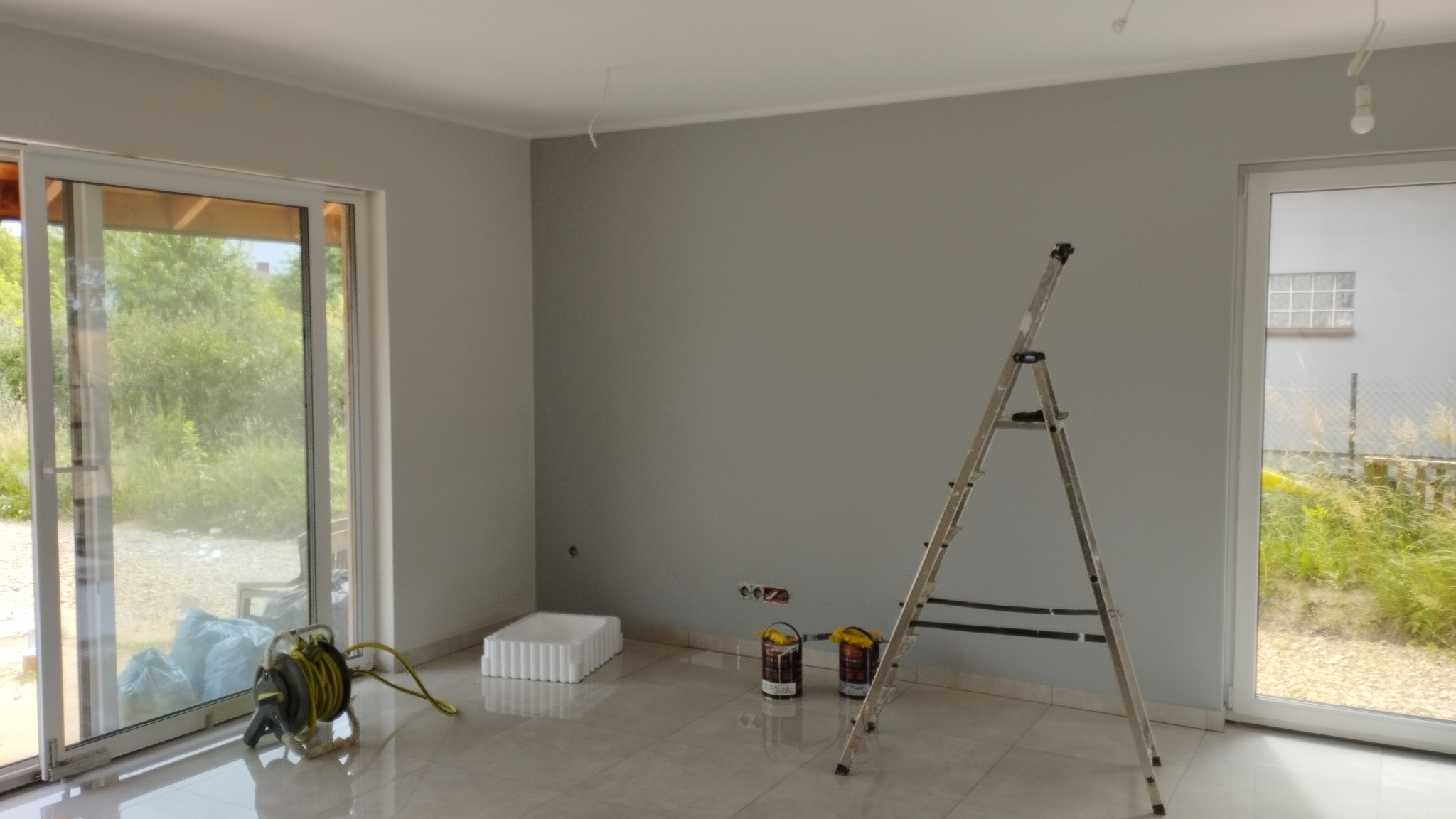 Wnętrze pokoju z pomalowanymi na szaro ścianami, drabina oparta o ścianę, dwie puszki z farbą na podłodze, zwinięty przedłużacz elektryczny i okno przesuwne z widokiem na zieleń.