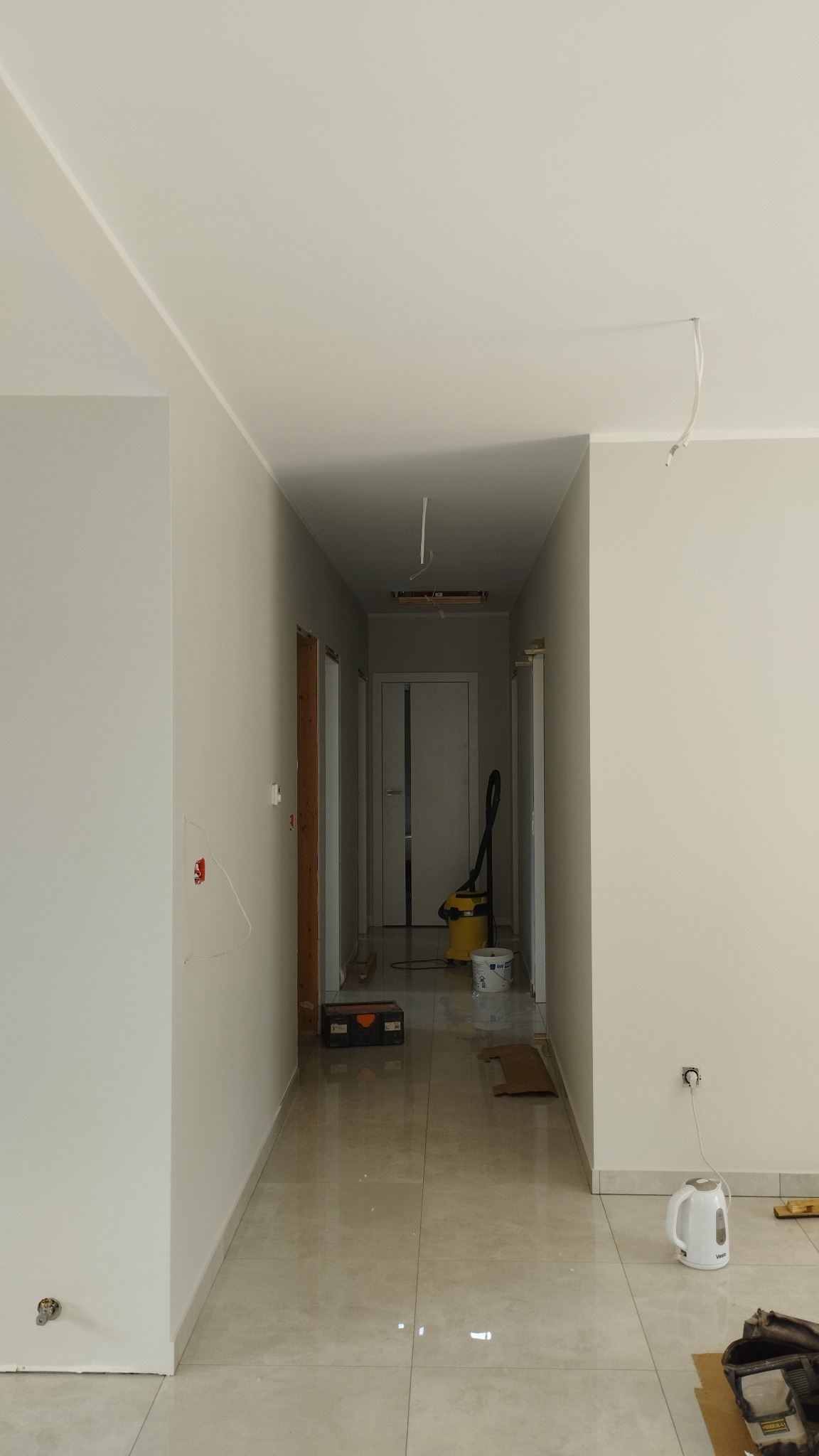 Korytarz w trakcie malowania, ściany w jasnych odcieniach szarości, widoczny żółty odkurzacz, biały czajnik elektryczny przy gniazdku i skrzynka z narzędziami na podłodze.