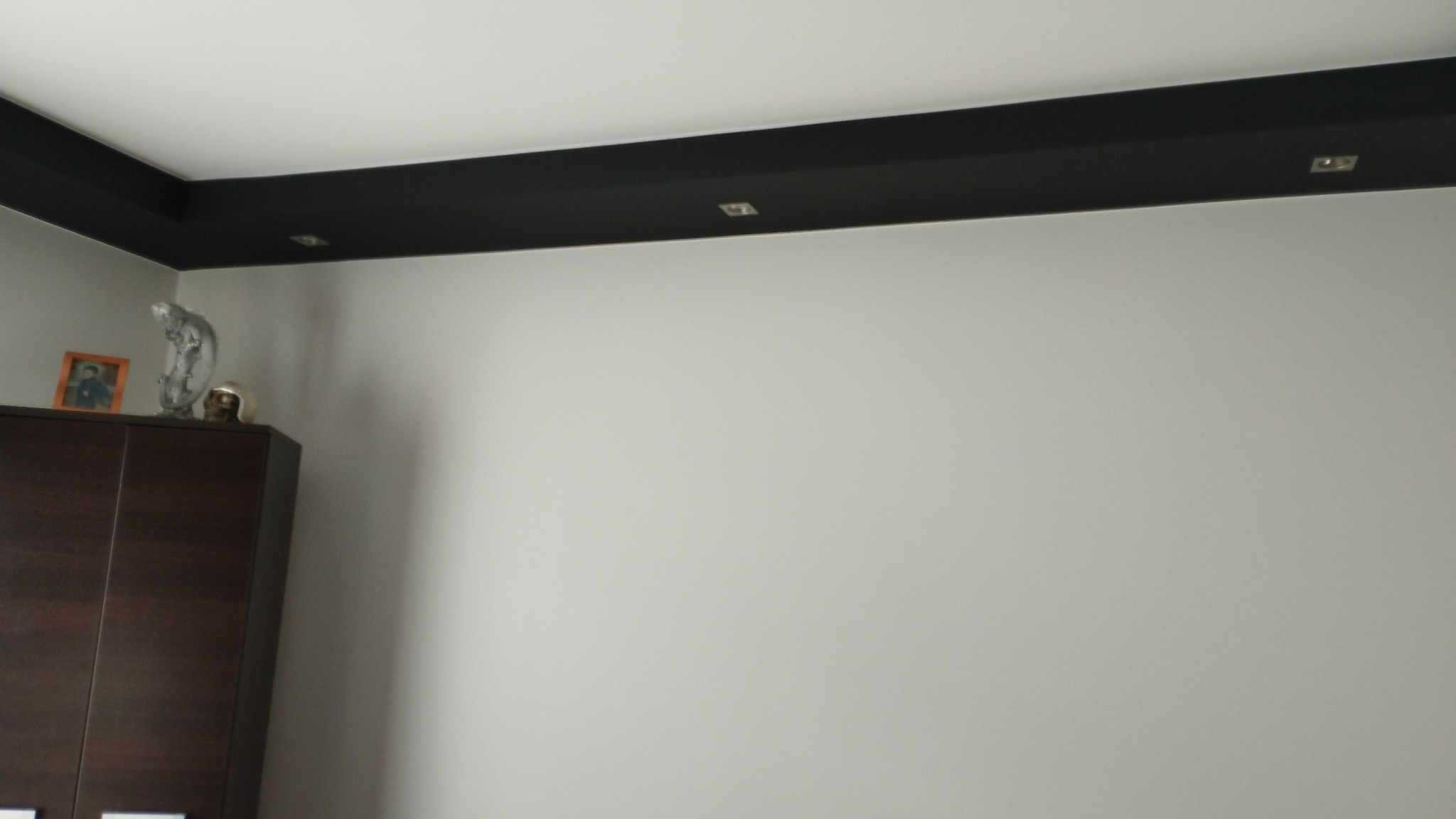 Pomieszczenie po malowaniu: szara ściana, czarny pas sufitowy z wbudowanymi punktami świetlnymi, ciemnobrązowa szafa z dekoracjami na górze.