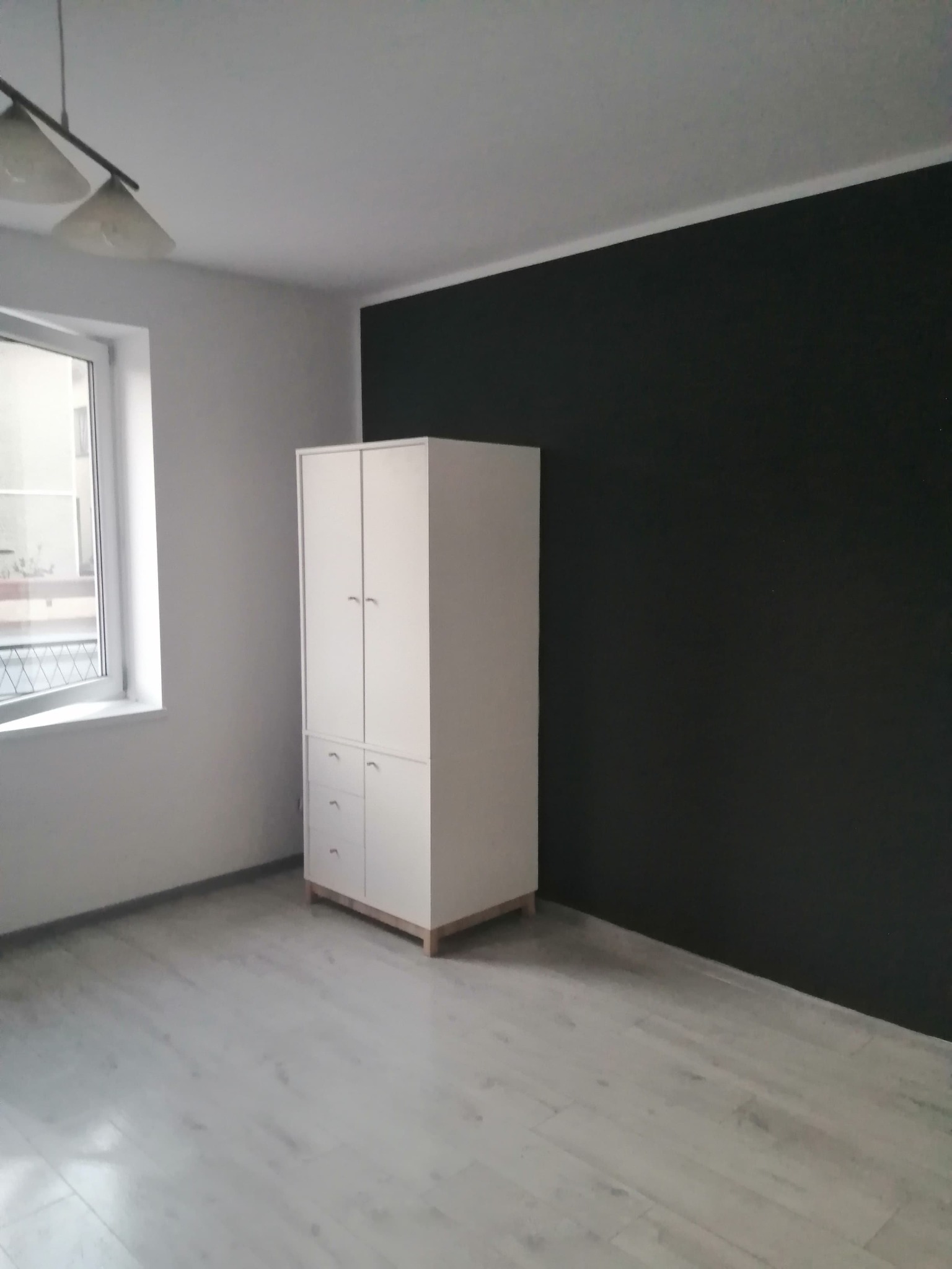 Pokój po odmalowaniu ścian: jedna ściana w kolorze grafitowym, pozostałe białe, jasna podłoga, biała szafa z szufladami, okno z widokiem na zewnątrz.