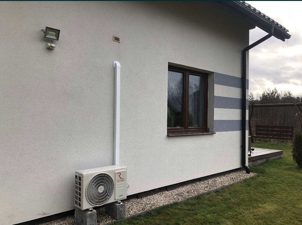 Biała jednostka zewnętrzna klimatyzatora marki Rotenso zamontowana na betonowych bloczkach przy ścianie domu z widoczną białą rurą odprowadzającą.