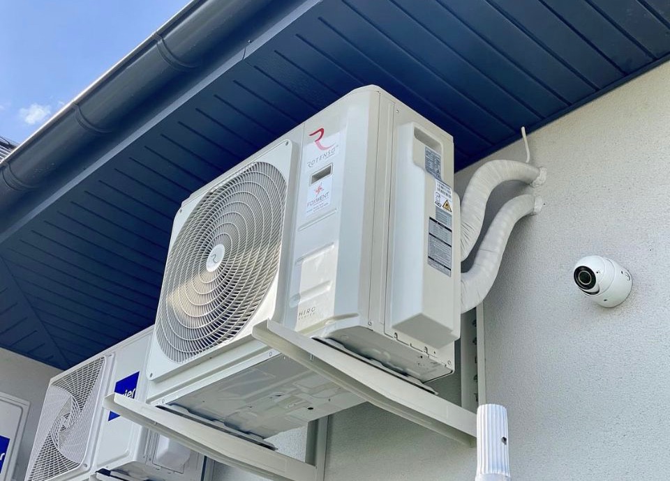 Biała jednostka zewnętrzna klimatyzacji Rotenso z widocznym logo i rurami w izolacji, zamontowana na jasnej elewacji budynku z kamerą monitoringu.