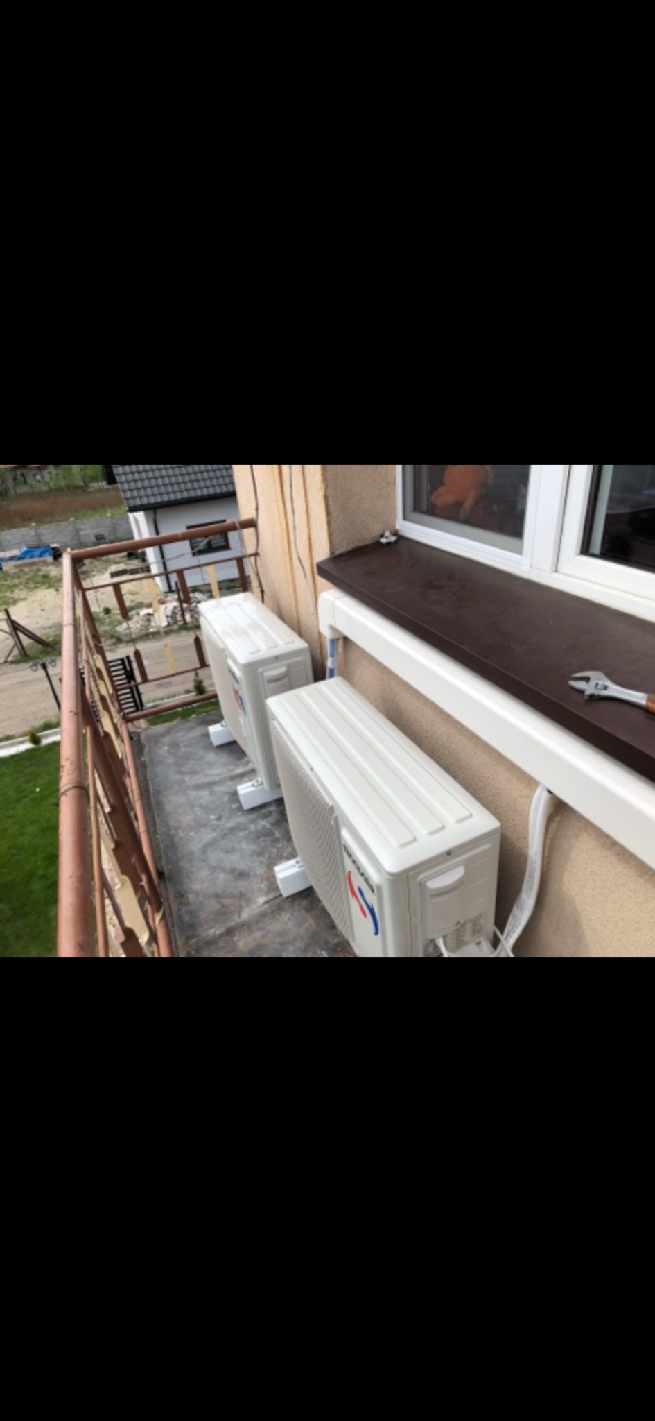 Dwa białe klimatyzatory zamontowane na balkonie, widoczny klucz nastawny leżący na parapecie, w tle dom z szarą dachówką i zielony trawnik.