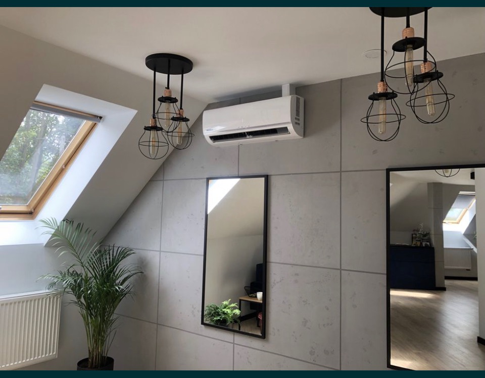 Wnętrze poddasza z zamontowanym, białym klimatyzatorem na ścianie z szarych paneli, widok na okno dachowe, lustro i lampy wiszące w stylu industrialnym.