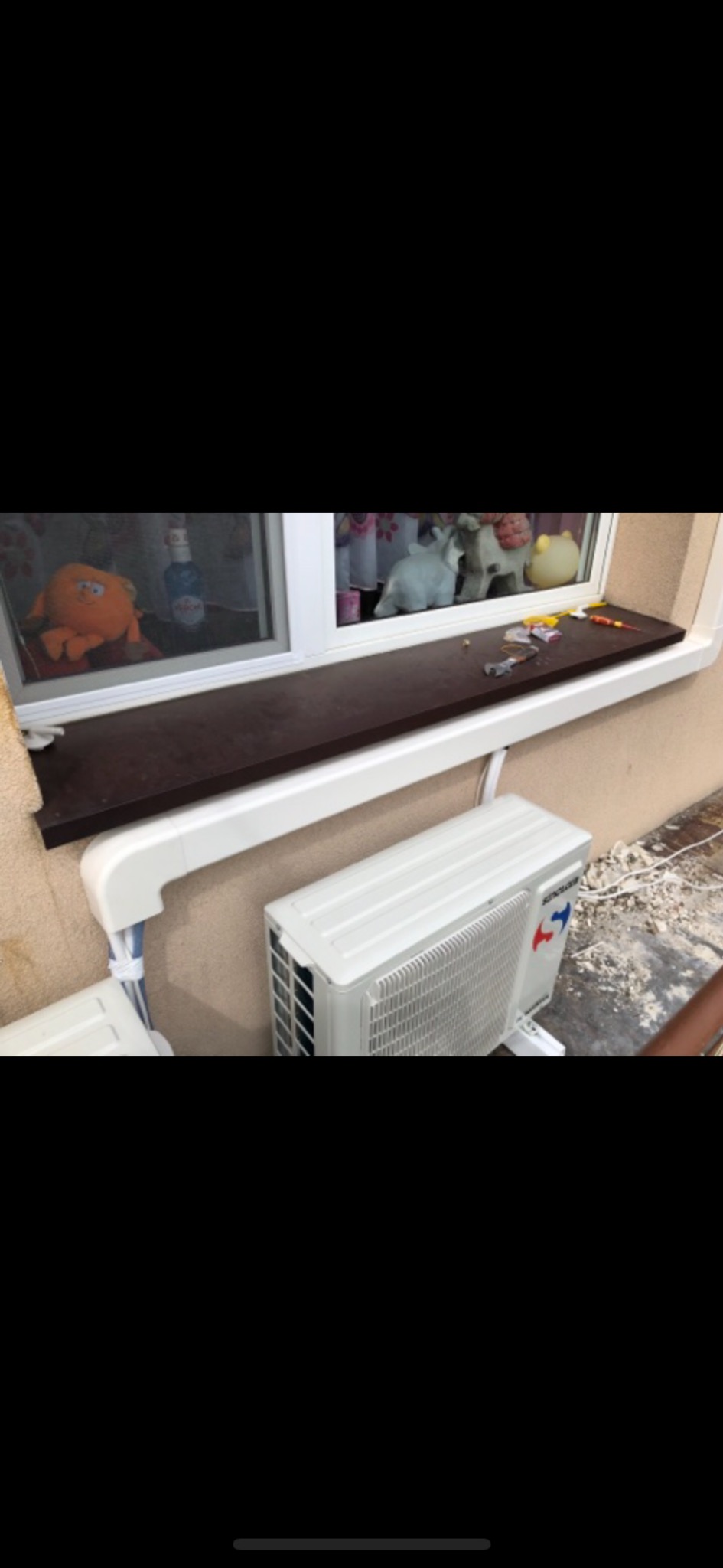Zewnętrzna jednostka klimatyzacji zamontowana pod oknem, widoczne przewody w białej listwie, na parapecie okiennym leżą narzędzia i drobne przedmioty.