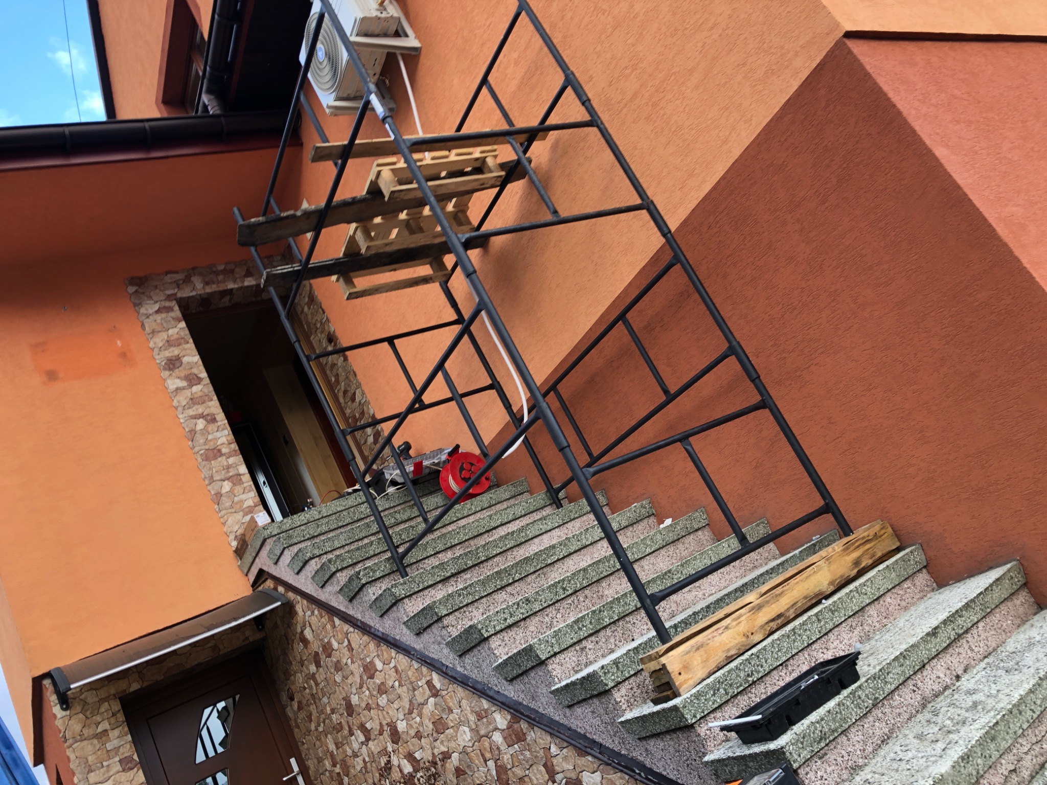 Montaż jednostki zewnętrznej na pomarańczowej ścianie domu, widoczne rusztowanie, schody wejściowe i narzędzia.