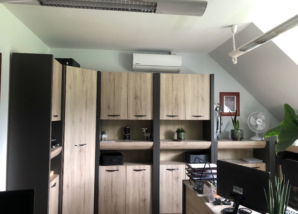 Biała jednostka klimatyzacyjna zamontowana nad drewnianymi szafkami w biurze na poddaszu, widoczny również wentylator i rośliny doniczkowe.