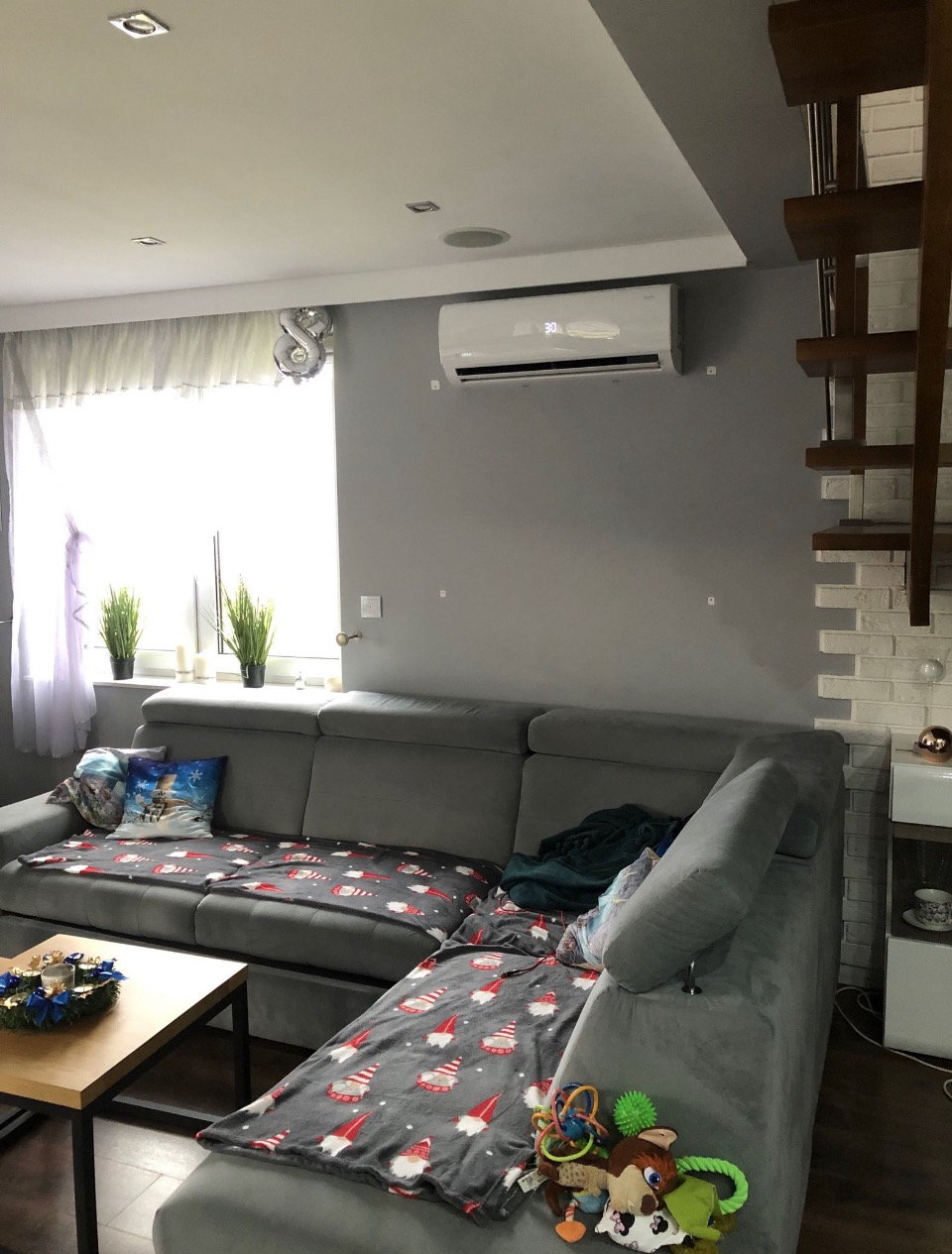 Biała jednostka klimatyzacyjna zamontowana na szarej ścianie w salonie nad szarą kanapą z dekoracyjnymi poduszkami i kocem w gnomowy wzór. Widoczna temperatura na wyświetlaczu klimatyzacji.