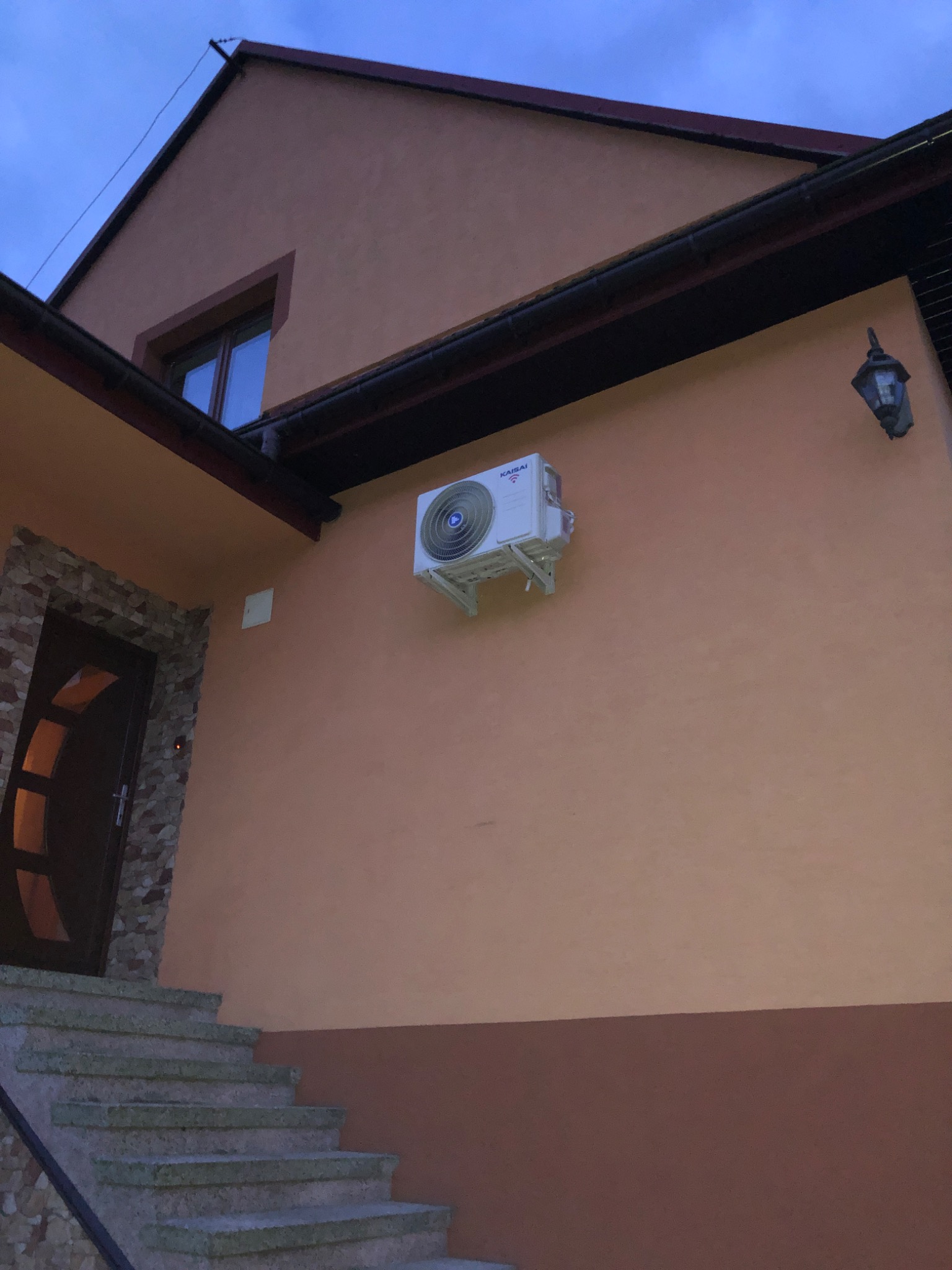Zewnętrzna jednostka klimatyzacji Kaisai zamontowana na pomarańczowej ścianie domu z widocznymi schodami wejściowymi i lampą zewnętrzną, sfotografowana przy zachodzącym słońcu.